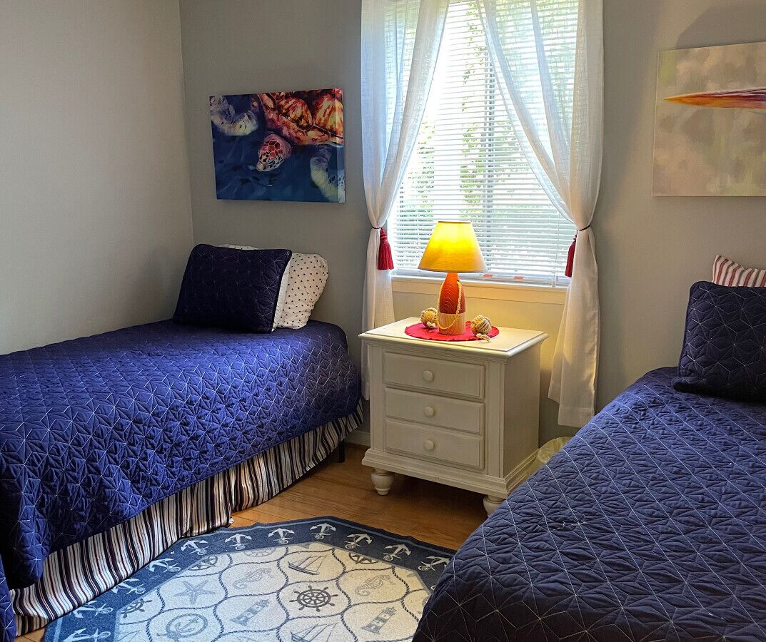 A bedroom with two beds, a nightstand with a lamp, a window with curtains, and a painting on the wall.