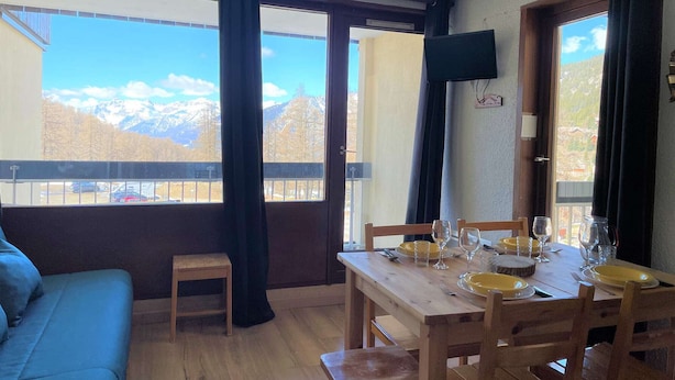 A room with a wooden dining table set for four, a blue sofa, and a view of snow-capped mountains through large windows.