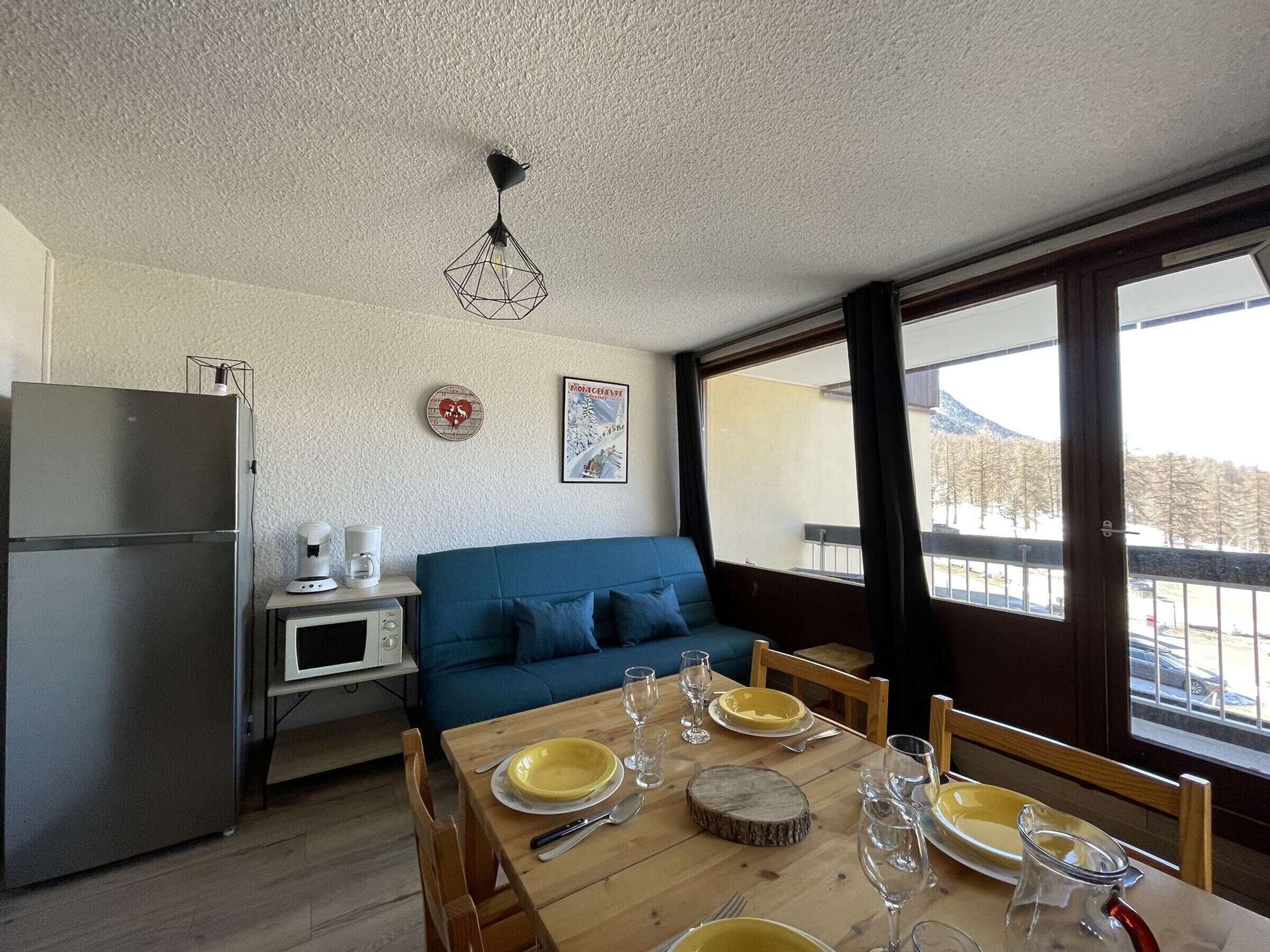 Ett kompakt bostadsutrymme med ett litet kök, ett matbord med plats för fyra personer, en blå soffa och ett stort fönster med utsikt över ett snötäckt landskap.