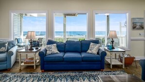A living room with a blue sofa, two side tables with lamps, a coffee table, and a view of the ocean through large windows. - "Sundance" - Oceanfront Home w/ Private Pool, 5 Bedrooms / 4.5 Bathrooms (Holden Beach)