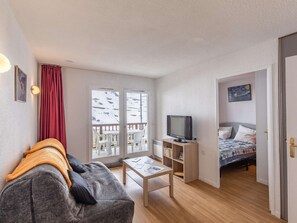 A compact room with a sofa, a television on a stand, a small table, and a bed with a patterned bedspread.