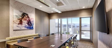 A conference room with a long table, chairs, a bench, a large screen, and a view through the glass doors.