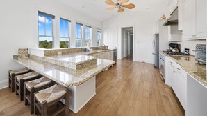 A spacious kitchen with granite countertops, wooden bar stools, and a view of the outdoors through large windows. - 🌺 Aloha Beaches Gulf-Front Retreat, Pool, Steps to Beach, Sleeps 14 (Panama City Beach)