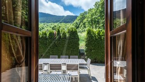 A view from inside a room with a large window overlooking a dining area with a table and chairs, and a forested mountain in the background. - Casa Eth Gauèc (Betrén)