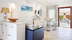 En moderne stue med sofa, lille bord, lampe, dekorativ sejlbåd, et billede, et køkkenområde og udsigt gennem en glasdør.