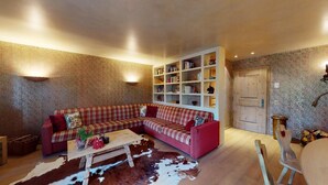 Un salon chaleureux avec un canapé à carreaux rouges, une table basse en bois et un tapis en peau de vache. On y voit une bibliothèque remplie de livres et une porte au fond.