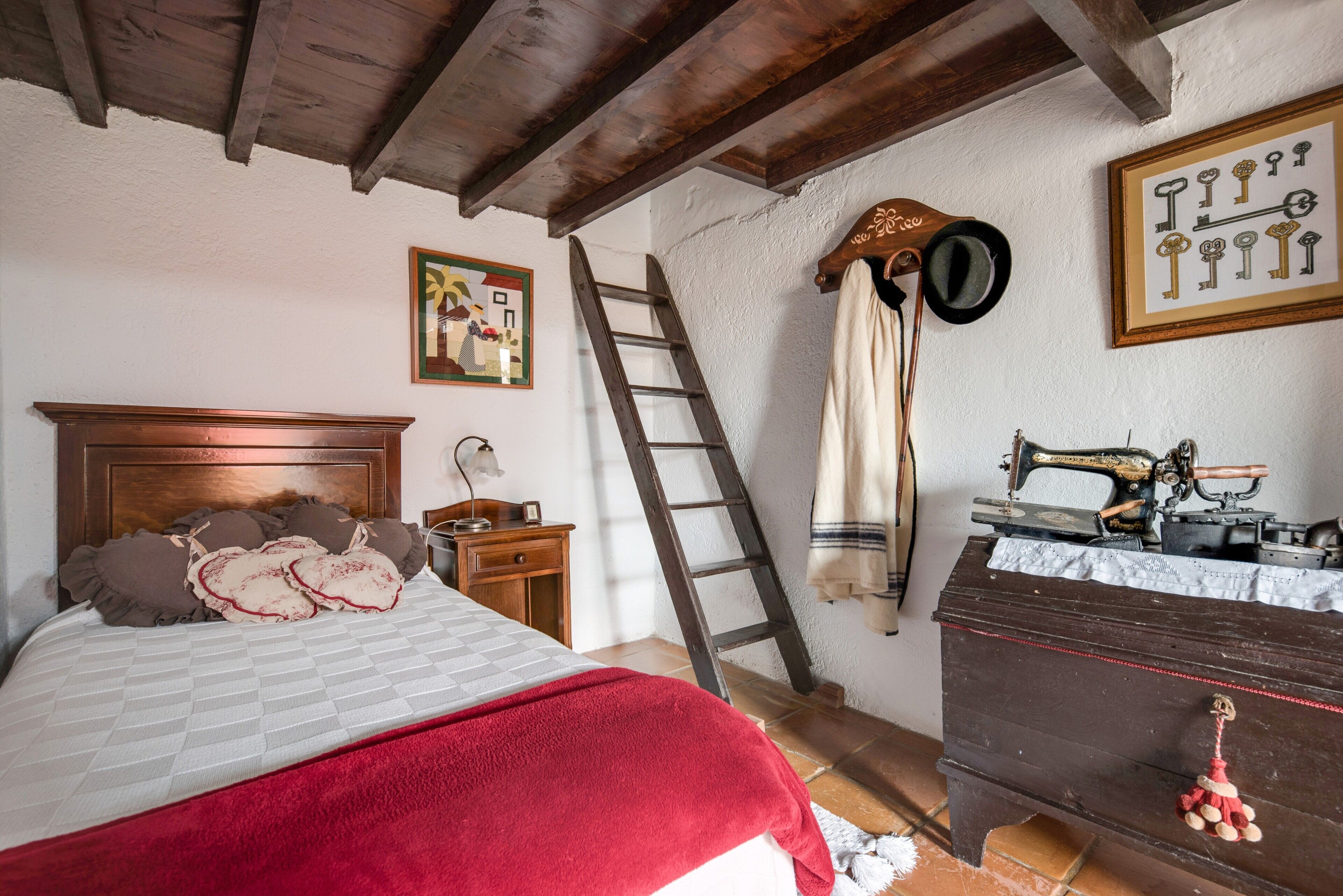 A bedroom with a wooden bed, a wooden dresser, a sewing machine, a ladder, and a wall-mounted hat rack.