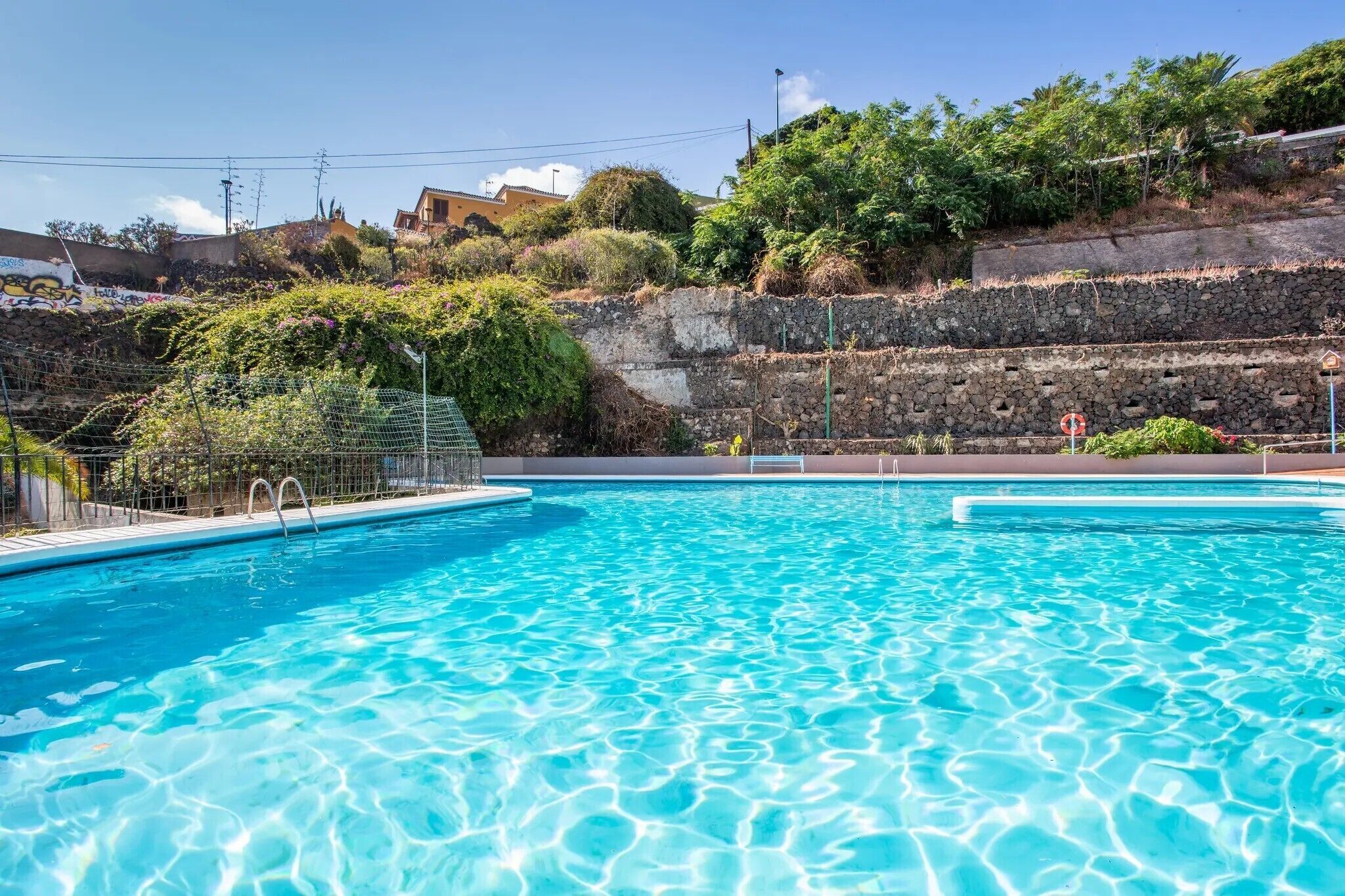 Uma piscina azul cristalina com parede de pedra e vegetação ao fundo.