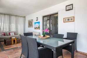 A dining area with a glass table and wicker chairs, a sofa with cushions, a small wooden side table, and a window with curtains.