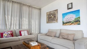 Una sala de estar con un sofá, dos mesas auxiliares, un cuadro y un ventanal con cortinas.