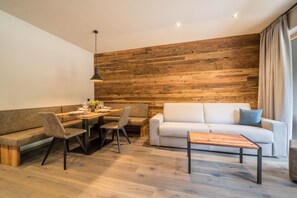 A modern living room with a wooden accent wall, a sofa, a dining table with chairs, and a coffee table. - Apartment “Seebrunn - Schusterhüttl Alm” with Mountain and Lake View, Terrace & Garden (Ulten)