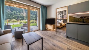 A modern living room with a TV, sofa, and coffee table, a kitchen with a sink, and a balcony with a view of a forested hillside. - Apartment “Seebrunn - "Äußere Schwemmalm” with Mountain View, Wi-Fi and Sauna (Ulten)