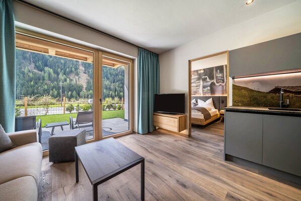 Un salon moderne avec un téléviseur, un canapé et une table basse, une cuisine avec un évier et un balcon offrant une vue sur une colline boisée.