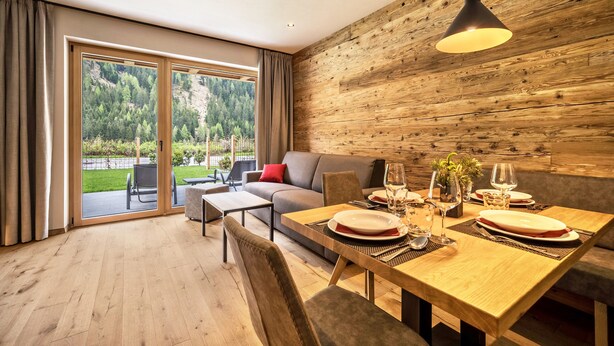 A modern dining area with a wooden table set for two, a sofa, and a view of a forested landscape through large sliding doors. - Apartment 'Seebrunn - Kuppelwieser Alm' with Mountain View, Private Terrace and Wi-Fi (Ulten)