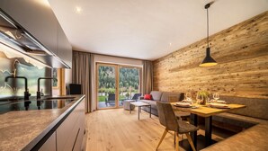 A modern kitchen with a central island, a dining area with a wooden wall, and a view of a green landscape through large windows. - Apartment 'Seebrunn - Kuppelwieser Alm' with Mountain View, Private Terrace and Wi-Fi (Ulten)