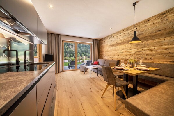 A modern kitchen with a central island, a dining area with a wooden wall, and a view of a green landscape through large windows.