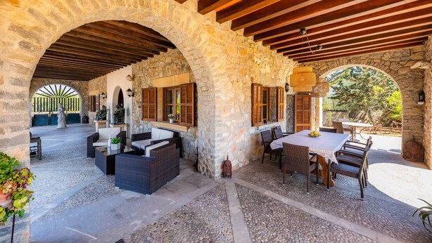 Patio esterno con archi in pietra, travi in legno, mobili in vimini e un tavolo con sedie.