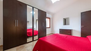 A bedroom with a large wardrobe, a bed with a red bedspread, a small bedside table, and a mirror on the wall. - Villa "Giulia Grande" in rural area near the sea with mountain view and garden (Castiadas)