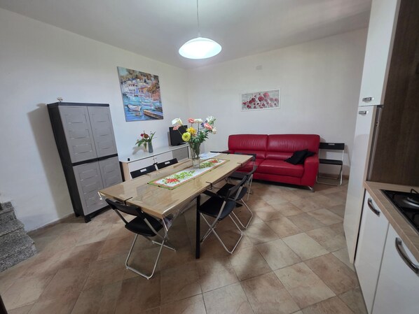 A compact living space with a dining table, chairs, a red sofa, a kitchen area with cabinets, and artwork on the walls. - Villa "Giulia Grande" in rural area near the sea with mountain view and garden (Castiadas)