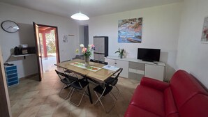 A dining area with a table, chairs, a vase of flowers, a clock, a painting, a television, and a red couch. - Villa "Giulia Grande" in rural area near the sea with mountain view and garden (Castiadas)