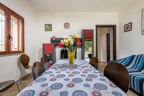 A dining area with a patterned tablecloth, chairs, a vase with flowers, and a clock on the wall.