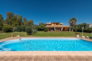 Una piscina con bordo piastrellato, una casa con tetto in tegole e vegetazione circostante.