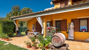 Une maison jaune avec un patio couvert, des plantes en pot, du mobilier d’extérieur et un tonneau sur lequel on peut lire « MP ».