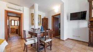 Un coin repas avec une table et des chaises en bois, un téléviseur fixé au mur et une cuisine visible à travers une porte ouverte.