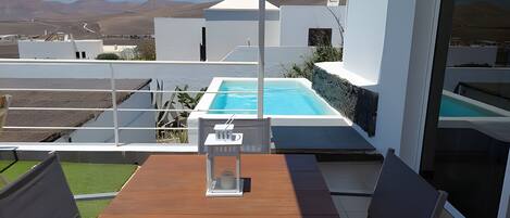 Un área de piscina moderna al aire libre con una mesa y sillas de madera, rodeada de paredes blancas y una vista despejada de las montañas.
