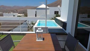 A modern outdoor pool area with a wooden table and chairs, surrounded by white walls and a clear view of the mountains. - Holiday Apartment “Amanecer ” with Ocean and Mountain View, Terrace and Pools (Tías)