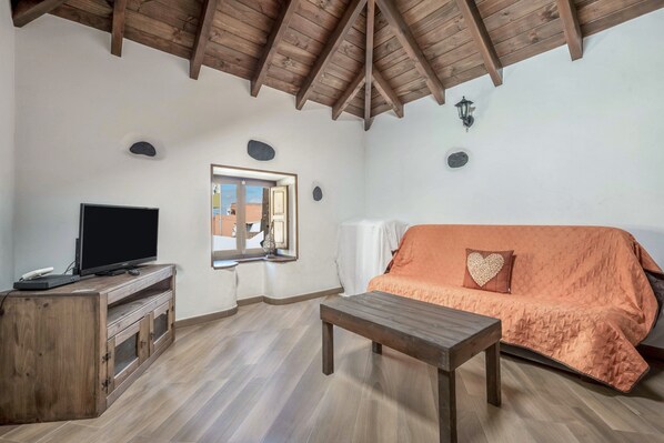 Una habitación con techo de madera, un televisor sobre un mueble de madera, un sofá con funda naranja y una ventana con vistas.