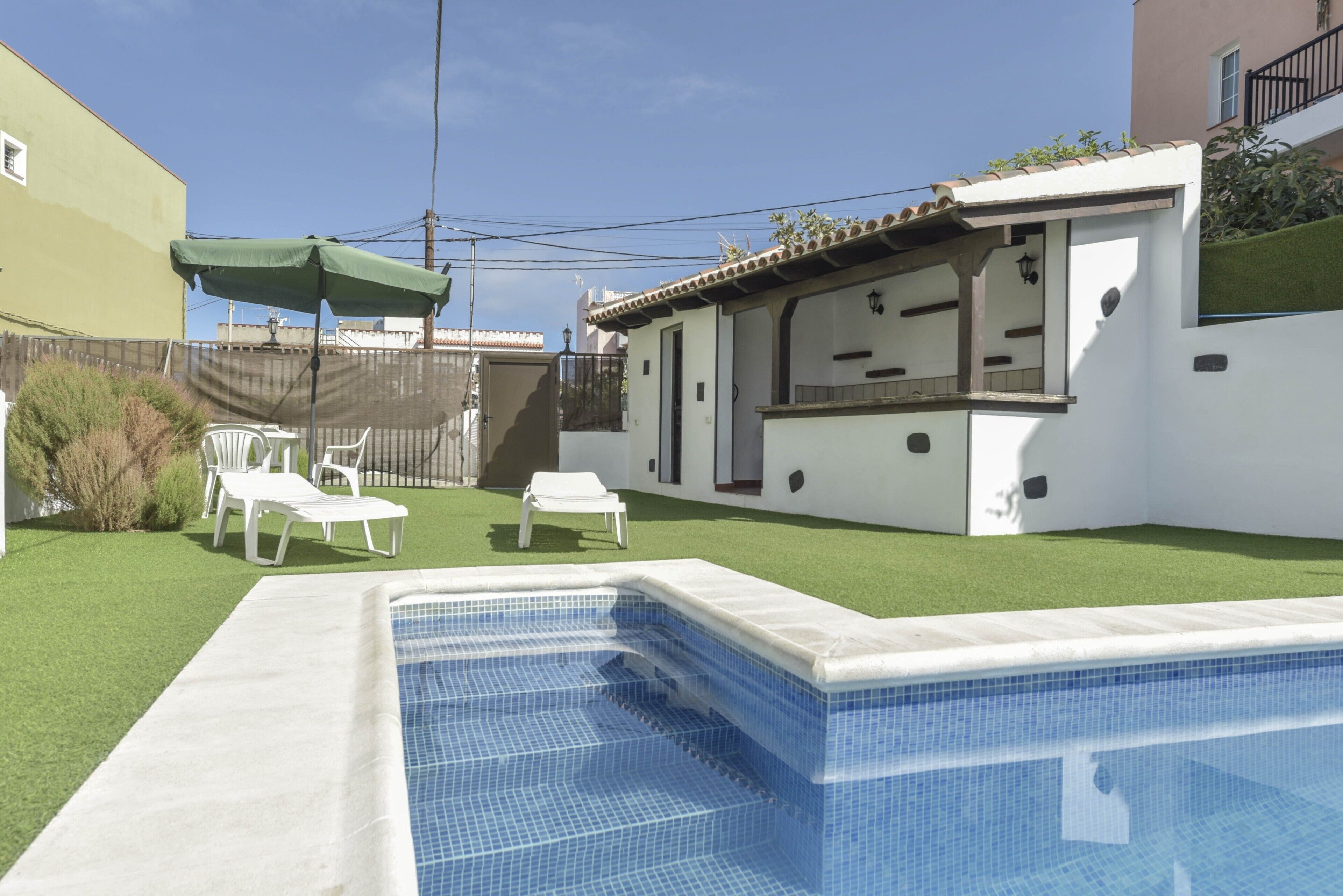 A swimming pool with a tiled edge, a green lawn, white lounge chairs, and a white building with a balcony.
