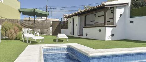 A swimming pool with a tiled edge, a green lawn, white lounge chairs, and a white building with a balcony.