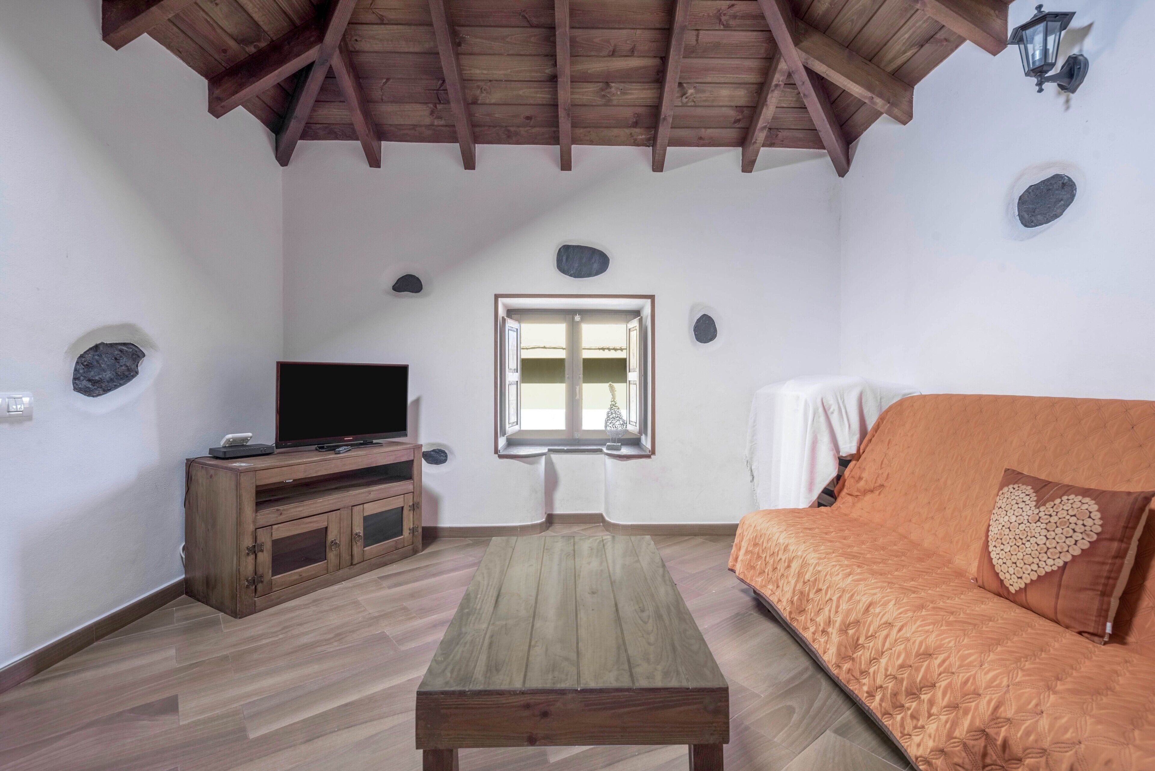 A room with a wooden ceiling, a television on a wooden cabinet, a sofa with a heart-shaped pillow, and a window with a view.