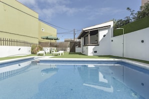 Una piscina con plataforma de baldosas, un pequeño patio con sillas y un edificio blanco con techo inclinado.