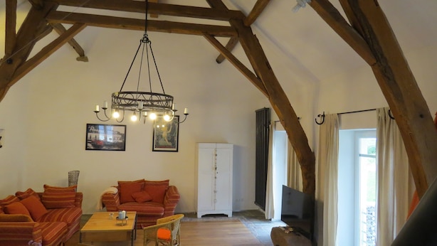 Un loft spacieux avec des poutres apparentes, un lustre, un ensemble de canapés, un meuble et un téléviseur.
