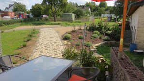 Un espace patio aménagé avec une table et des chaises, un chemin en pierre et un jardin où poussent différentes plantes, ainsi qu’un petit abri de jardin.