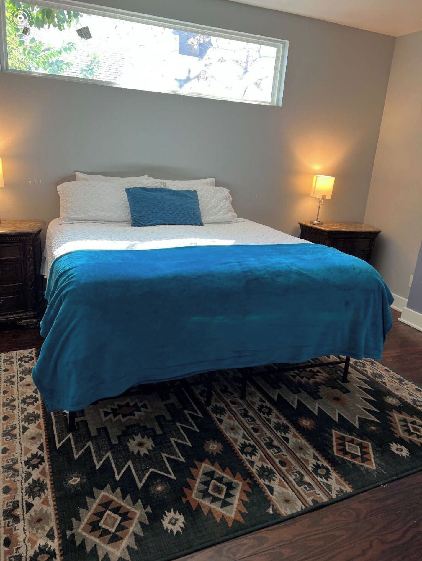 Un lit bien fait, avec une couvre-lit bleue et des oreillers blancs, un tapis à motifs et deux lampes de chevet posées sur des tables de nuit en bois foncé.