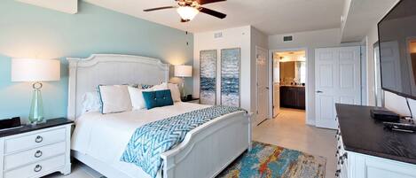 A bedroom with a bed, nightstands, a ceiling fan, and a view into the adjacent kitchen.