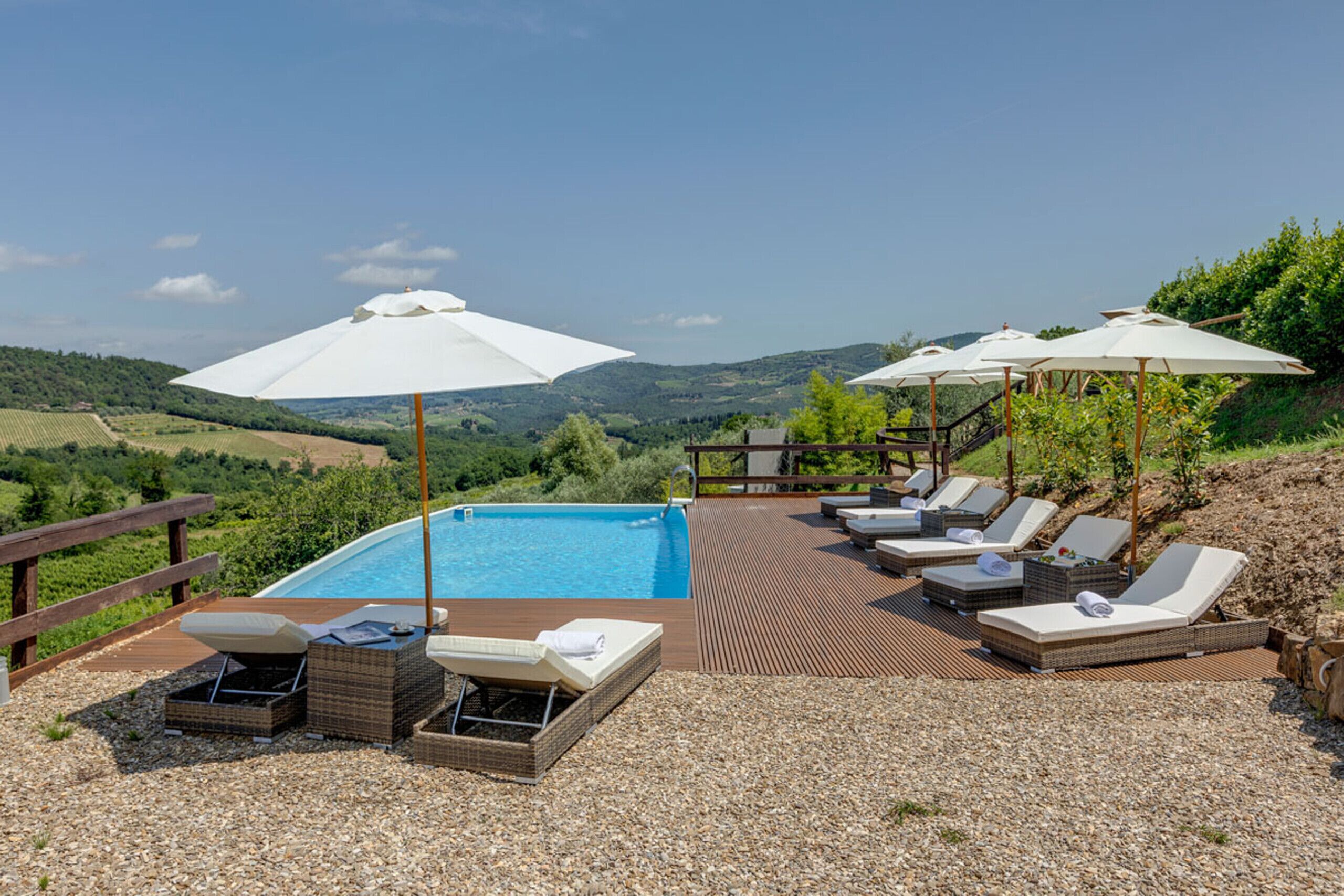 A poolside area with lounge chairs and umbrellas, surrounded by a wooden deck and gravel, overlooking a hilly landscape.