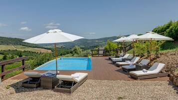 A poolside area with lounge chairs and umbrellas, surrounded by a wooden deck and gravel, overlooking a hilly landscape.