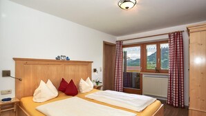 A bedroom with two beds, a wooden headboard, a nightstand, a window with curtains, and a view of mountains. - Ferienwohnung Jenner, 2-4 Personen, Infrarotkabine, 65 qm, Balkon mit Bergblick (Schönau am Königssee)
