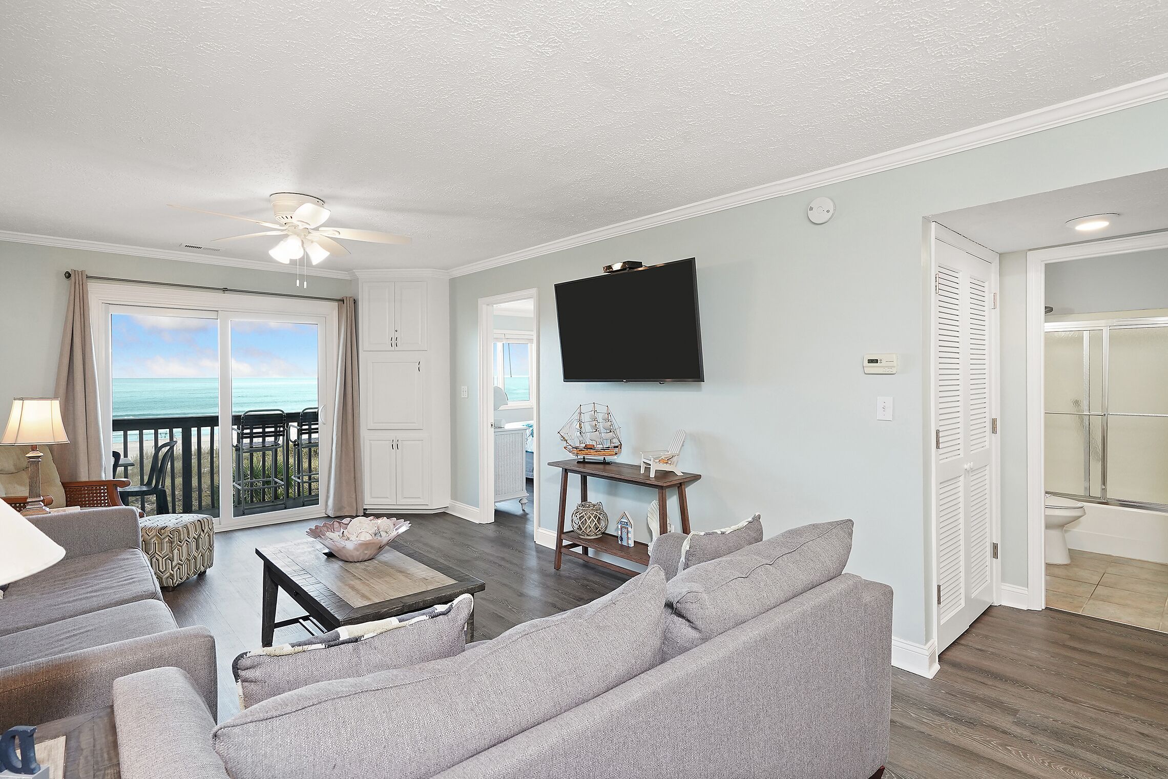 A living room with a view of the ocean, a ceiling fan, a flat-screen TV, and a dining area.