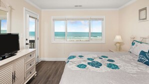 A bedroom with a large window offering a view of the ocean, a television on a dresser, and a bed with floral bedding. - Condo w/ Private Balcony & Stunning Beach Views (Surfside Beach)