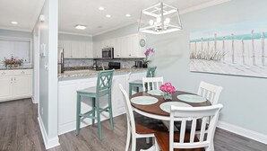 A modern kitchen with white cabinets, a central island, and a dining area with a round table and chairs. - Condo w/ Private Balcony & Stunning Beach Views (Surfside Beach)