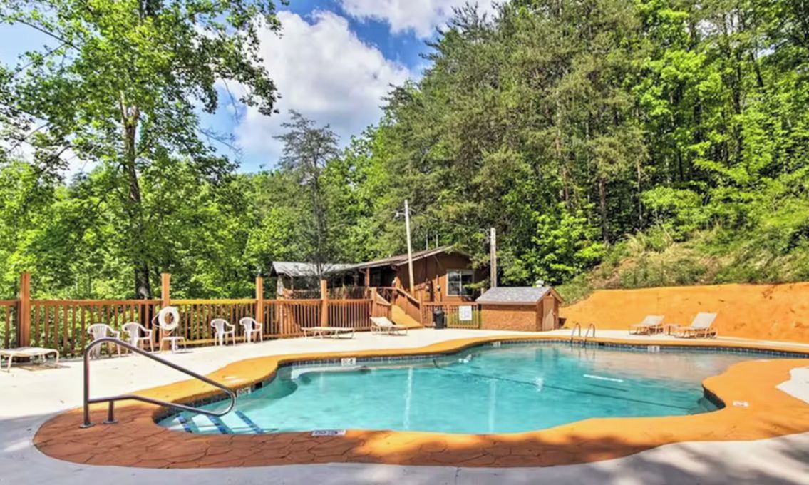 A swimming pool with a wooden deck, surrounded by trees and a small building with a roof.