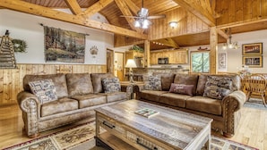 Interior rústico de una cabaña con vigas de madera, un techo de madera grande, un sofá para dos personas, una mesa de centro y una zona de cocina.