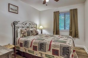 Ein Schlafzimmer mit einem Bett, einem Nachttisch, einer Lampe, einem Fenster mit Vorhängen und einem an der Wand hängenden Bild.