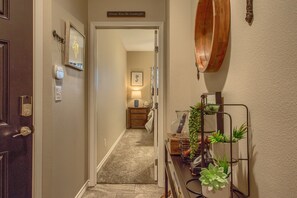 A hallway with a door on the left, a sign above the door, a light fixture, a mirror, and a plant stand.