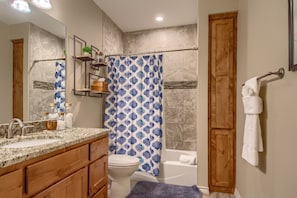 A bathroom with a marble sink, wooden cabinets, a toilet, a shower with a blue patterned curtain, and a wooden door.